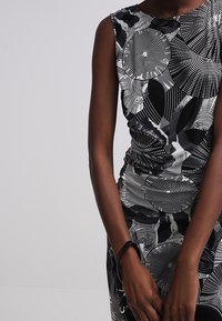 Ärmlös klänning med svarta och vita abstrakta cirkulära och linjemönster, bärs av en person mot en enfärgad bakgrund.