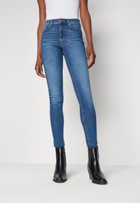 Jeans ajustados de denim azul con una cintura de media altura. Incluye un cierre frontal con botón, cinco bolsillos y un corte slim a lo largo de las piernas.
