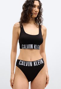 Γυναίκα που φοράει μαύρο bralette Calvin Klein και αντιστοιχούσες σλιπ με ψηλό κόψιμο και λευκές κορδέλες με λογότυπο, στέκεται μπροστά σε λευκό φόντο.