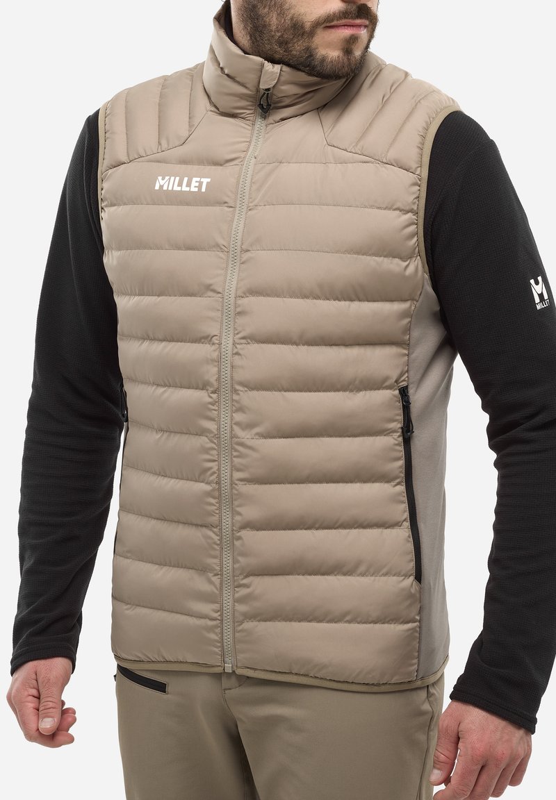 Beige gepolsterte Weste mit hohem Kragen, Frontreißverschluss und schwarzen langen Ärmeln. Verfügt über Seitentaschen und ein Logo auf der Brust. Glatte Textur.