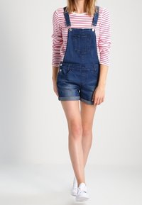 Denim short overalls with adjustable straps, front pocket, and side button details. Worn over a red and white striped long-sleeve shirt.