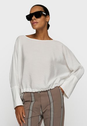 Kobieta w oversize'owej białej teksturowanej bluzce, brązowych spodniach z czarno-białymi pionowymi paskami oraz dużych czarnych okularach przeciwsłonecznych, pozująca z ręką w kieszeni.