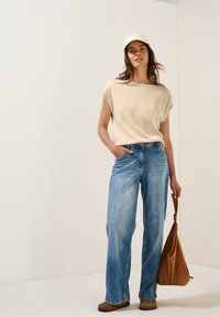 Beige kortärmad stickad topp, löst sittande blå vida jeans, bruna mocka anklarboots och en beige keps. En brun mockaväska hänger från handen.