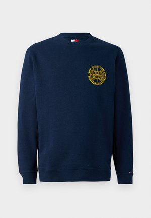 Tummansininen crewneck-huppari, jossa on keltainen "Tommy Jeans New York City" -logo vasemmalla rinnassa ja pieni lippu vasemmassa hihassa.