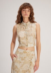 Blusa sin mangas cortada en tela beige claro con patrón floral crema; cuenta con cierre de botones y un pequeño cuello, textura suave.