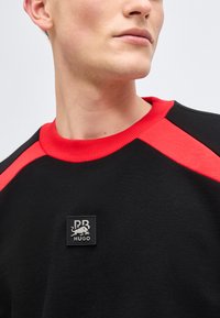 Νεαρός άνδρας που φοράει μαύρο φούτερ με κόκκινο γιακά και λεπτομέρειες στους ώμους, με ένα τετράγωνο λογότυπο "PB HUGO" στο στήθος.