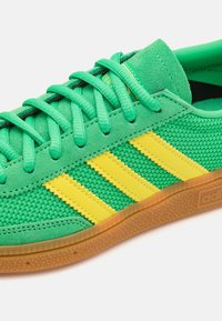 Πράσινα αθλητικά παπούτσια με διχτυωτό και σουέτ υλικό, διαθέτουν τρεις κίτρινες ρίγες, υφή από καουτσούκ στο πέλμα και ίσιες πράσινες κορδόνες.