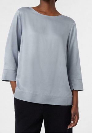 Femme portant un chemisier gris clair à manches trois-quarts assorti d’un pantalon noir, debout avec une main dans sa poche.