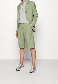 Blazer verde claro y pantalones cortos a juego, hechos de una tela suave, combinados con un suéter gris con cremallera y zapatillas deportivas plateadas.