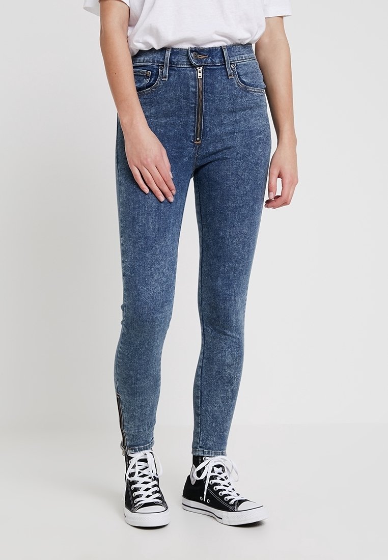 Jean en denim taille haute avec une fermeture éclair frontale, coupe slim, délavage bleu clair, et détail de fermeture éclair sur le côté. Associé à des baskets montantes noires.