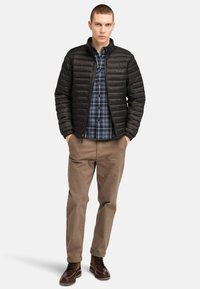 Giacca imbottita nera con zip frontale, indossata sopra una camicia a quadri blu. Abbinata a pantaloni beige e stivali marroni con lacci. Sfondo neutro.