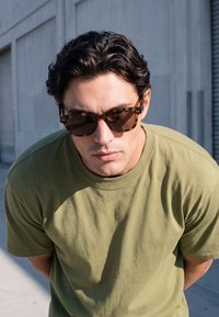 Ung mand iført skildpaddemønstrede solbriller og en olivengrøn t-shirt, der læner sig let fremad udendørs nær en grå bygning.