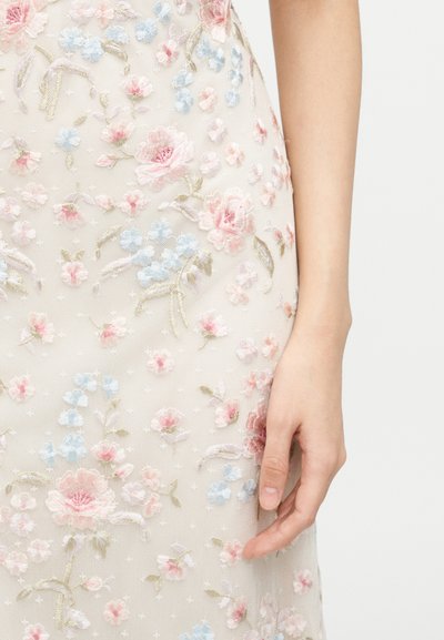 Κοντινό πλάνο ενός χεριού που ακουμπά δίπλα σε ένα κρεμ φόρεμα με λεπτή ροζ, μπλε και πράσινη κεντητή ανθολογία.