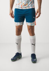 Conjunto de fútbol de Puma que incluye una camiseta blanca con un diseño, pantalones cortos azules y calcetines blancos con un logo negro. Las zapatillas de fútbol son de color azul oscuro con acentos verdes.