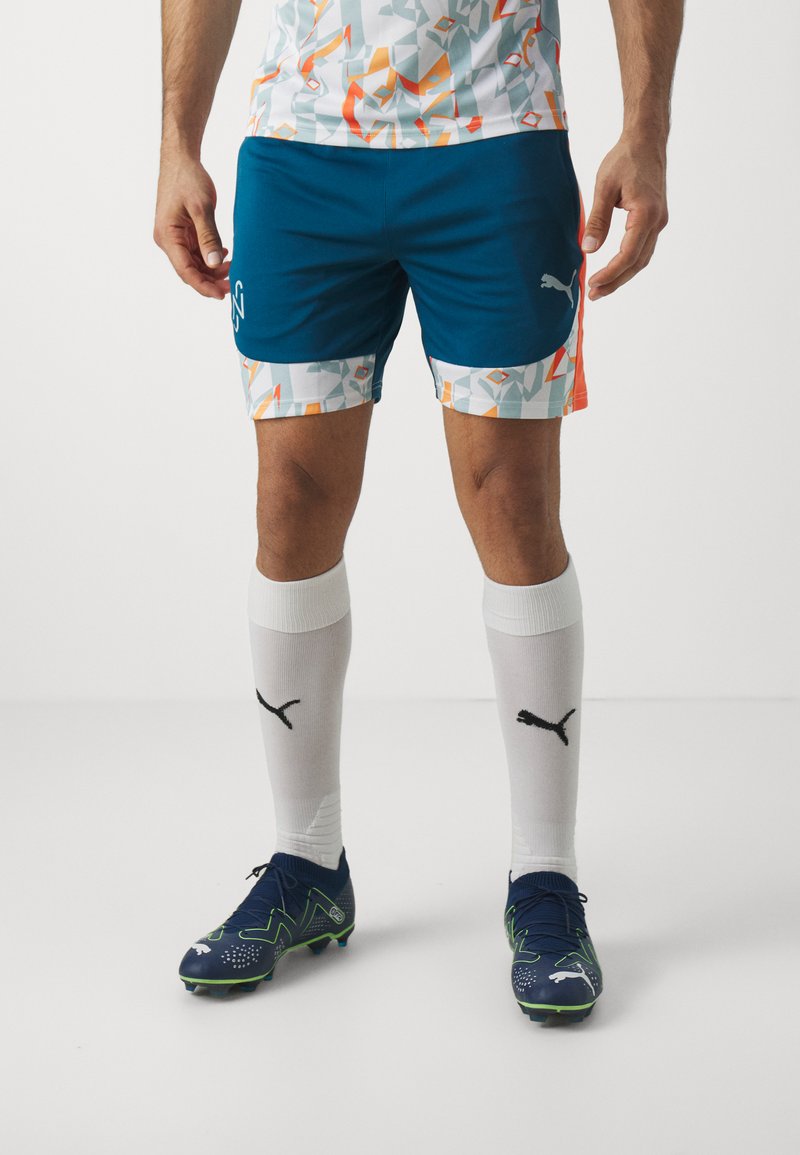 Conjunto de fútbol de Puma que incluye una camiseta blanca con un diseño, pantalones cortos azules y calcetines blancos con un logo negro. Las zapatillas de fútbol son de color azul oscuro con acentos verdes.