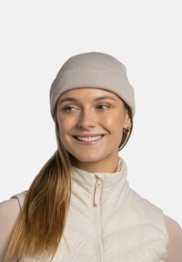 Light beige knitted beanie with a ribbed texture, worn with a cream sleeveless puffer vest. The beanie is snug-fitting and simple in design.