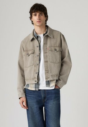 Un hombre luce una chaqueta beige abotonada sobre una camisa de cuadros y una camiseta blanca, combinada con unos vaqueros azul oscuro, sobre un fondo sencillo de estudio.