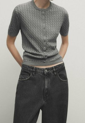 Woman wearing a grey short-sleeve cable knit buttoned top tucked into high-waisted black denim jeans with hands in pockets.