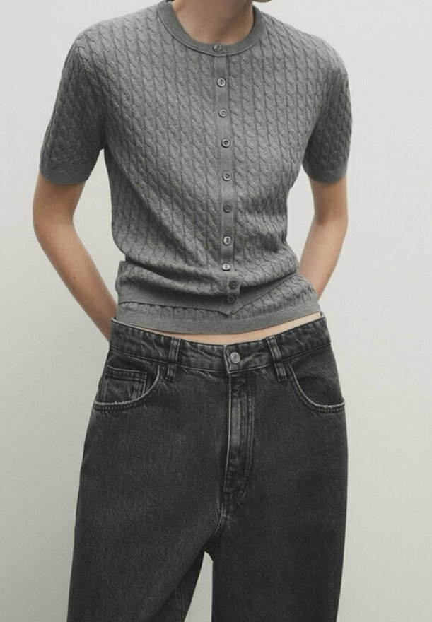 Femme portant un haut gris à manches courtes en maille torsadée avec boutons, rentré dans un jean en denim noir taille haute, les mains dans les poches.