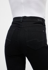 Jeans negros de cintura alta, con dos bolsillos traseros, detalles de costura sutil y una textura de tela lisa y elástica.