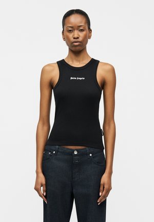 Γυναίκα που φοράει μαύρο αμάνικο ριμπ τοπ με το κείμενο «Palm Angels», συνδυασμένο με σκούρα μπλε τζιν, στέκεται μπροστά σε έναν απλό φόντο.