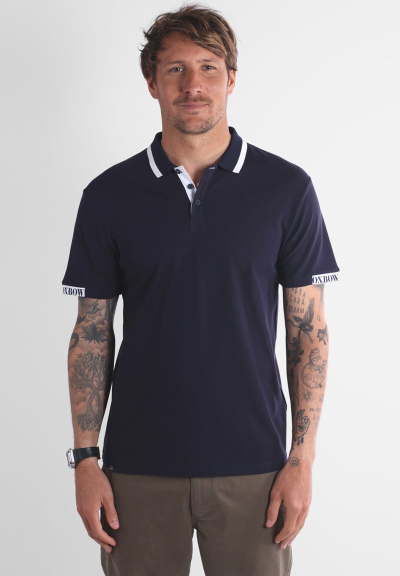 Homme avec des tatouages sur les deux bras portant un polo bleu marine avec des finitions blanches et un pantalon marron, se tenant devant un fond uni.