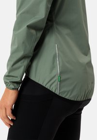 Lichte groene jas met elastische manchetten, reflecterende zijrits en een zwarte textuur onderrand, gecombineerd met zwarte leggings.