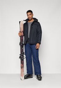 Skiutstyr som inneholder en mørk jakke, grå skjorte, blå bukser og svarte skistøvler, holding flerfargede ski med bindinger.