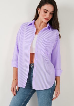 Naine kannab valge lõikega valgecrop topi peal helelilla ja valge triibulise nööpidega särgi ning siniste teksade ja kuldehetega.