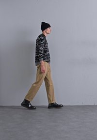 Suéter negro con patrones y acentos grises, pantalones beige y zapatos negros. El modelo lleva un gorro negro mientras camina frente a un fondo gris.