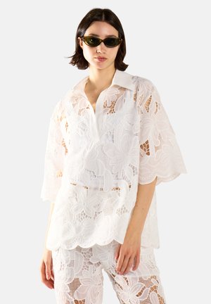 Kobieta ubrana w biały kwiatowy koronkowy strój z falistymi brzegami i ciemnymi owalnymi okularami przeciwsłonecznymi, stojąca na jednolitym jasnym tle.