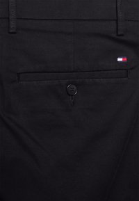 Schwarze Hose mit einer Gesäßtasche mit Knopfverschluss und einem kleinen rot-weiß-blauen Logo auf der rechten Seite. Baumwollmaterial.