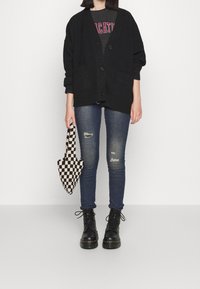 Svart överdimensionerad cardigan med knapplåsingar, bärs över en grafisk t-shirt. Slim-fit blå jeans med rivna detaljer. Svarta snörboots och en rutigt mönstrad väska.