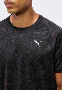 Κοντινό πλάνο ενός ατόμου που φοράει μαύρο t-shirt Puma με καμουφλάζ σχέδιο και λευκό λογότυπο Puma στο αριστερό μέρος του στήθους.