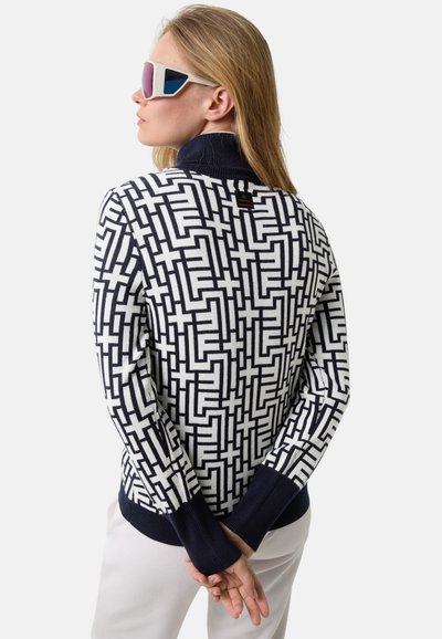 Kobieta z blond włosami, ubrana w sweter w białe i granatowe wzory geometryczne, białe spodnie i futurystyczne białe okulary przeciwsłoneczne, z twarzą zwróconą w lewo, z rękami za plecami.