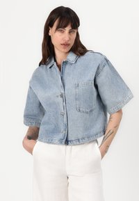 Giacca in denim blu tagliata con maniche corte, chiusura a bottoni e taschino sul petto. Lavaggio chiaro con una texture sbiadita.