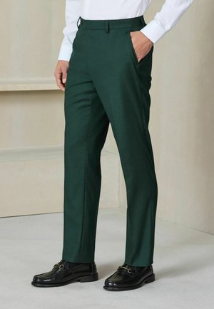Uomo che indossa pantaloni su misura verde scuro con camicia bianca e mocassini in pelle nera, in piedi con una mano in tasca.