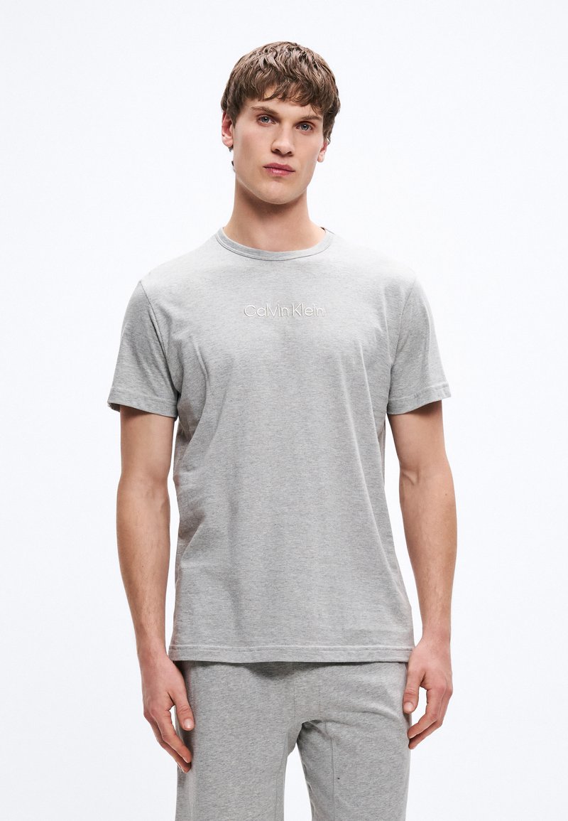 Hall hall puuvillast lühikeste varrukatega T-särk, ümara kaelusega ja rinnal tikitud "Calvin Klein" logo. Modell kannab sobivaid halli värvi lühikesi pükse.