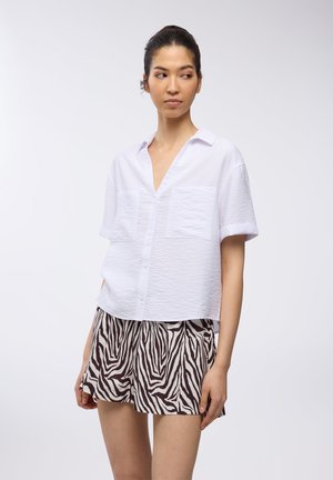 Camicia bianca a maniche corte con scollatura a V e due tasche frontali, abbinata a pantaloni corti a righe marroni e crema con un motivo a zebra.