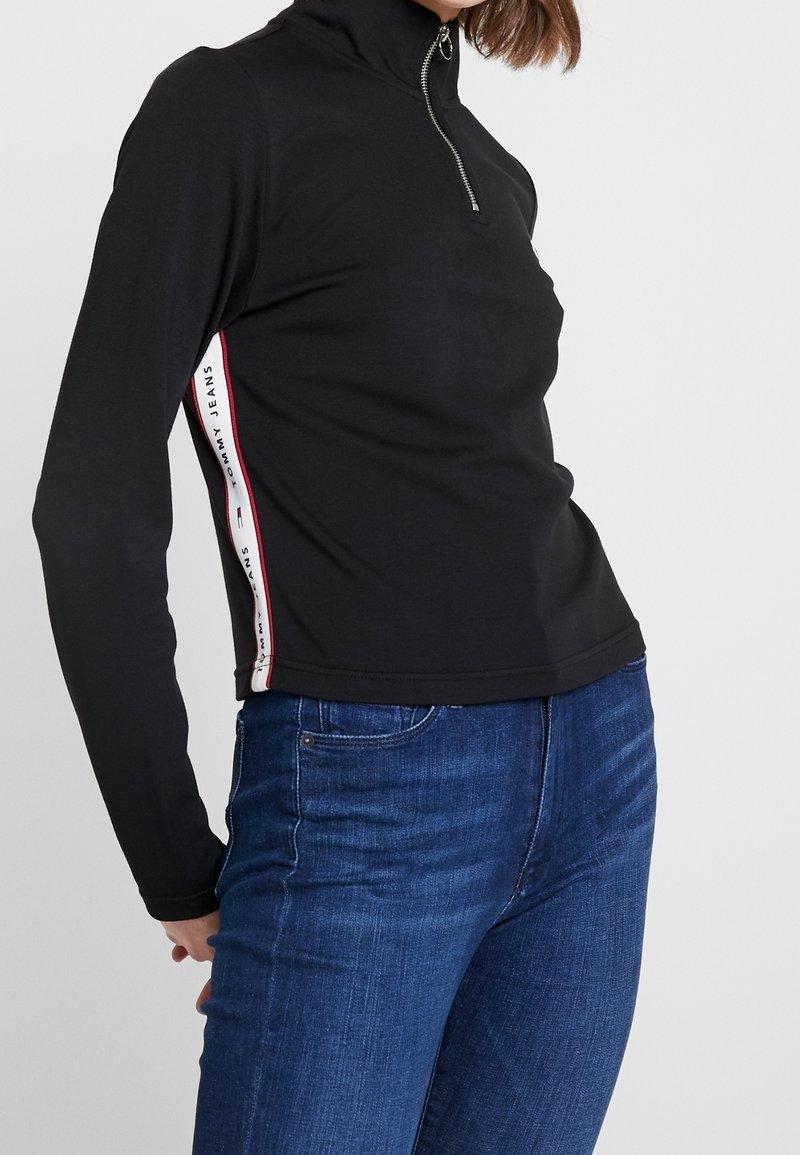 Haut noir à manches longues avec un col haut et une fermeture éclair à l'avant. Présente des bandes latérales blanches et rouges avec le texte "TOMMY JEANS". Tissu lisse, design ajusté.
