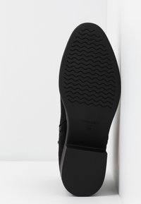 Semelle de chaussure noire avec un motif de bande de roulement ondulé, étiquetée "ANNA FIELD" et taille 38, positionnée contre un fond blanc en coin.