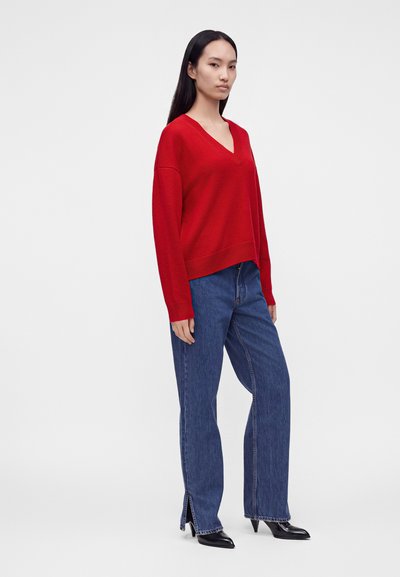 Femme debout portant un pull rouge à col en V, un jean bleu droit avec des fentes latérales, et des chaussures à talons pointus noires sur fond blanc.