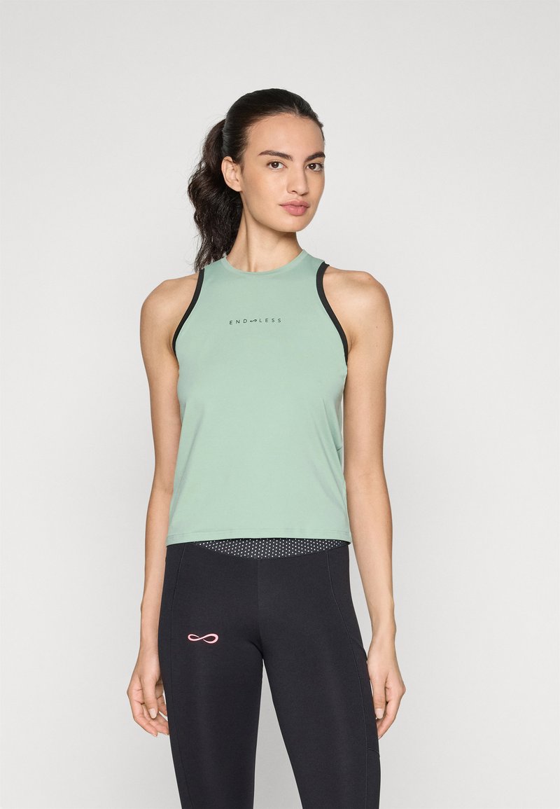Woman with dark hair in a ponytail wearing a sleeveless green top and black leggings, standing against a white background.