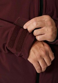 Burgunderfarbener Jackenärmel mit zwei Klettverschlussriemen. Nahaufnahme von Händen, die den Riemen über den glatten Stoff anpassen. Sichtbares Detail des Reißverschlusses auf der Vorderseite.