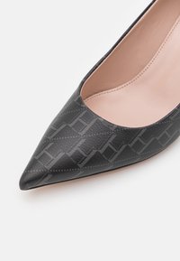 Escarpin à bout pointu en cuir noir présentant un motif texturé avec un logo discret, et une semelle intérieure lisse de couleur neutre.