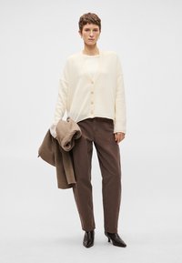 Γυναίκα που φοράει κρεμ καπιτονέ ζακέτα με κουμπιά, λευκό μπλουζάκι, καφέ παντελόνι, μαύρες στιλέτto μπότες, κρατώντας ένα καφέ παλτό, με φόντο απλή επιφάνεια.