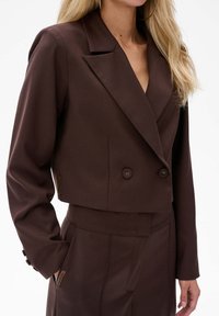 Brązowa krótka marynarka z gładkiego materiału, z podwójnym zapięciem, spiczastymi klapami i guzikami na rękawach.