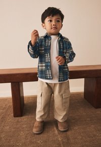 Barn iført en blå og grøn ternet flannel-skjorte, hvid T-shirt, beige cargobukser og brune ruskindsstøvler, stående ved en træbænk.