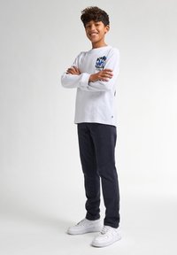 Witte longsleeve shirt met blauw grafisch ontwerp, gecombineerd met donkerblauwe broek en witte sneakers, met een relaxed fit en ronde hals.