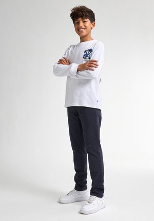 Witte longsleeve shirt met blauw grafisch ontwerp, gecombineerd met donkerblauwe broek en witte sneakers, met een relaxed fit en ronde hals.